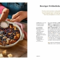 Hands dusting a berry breakfast casserole with powdered sugar over a round baking dish on a wooden table, with cubed bread nearby