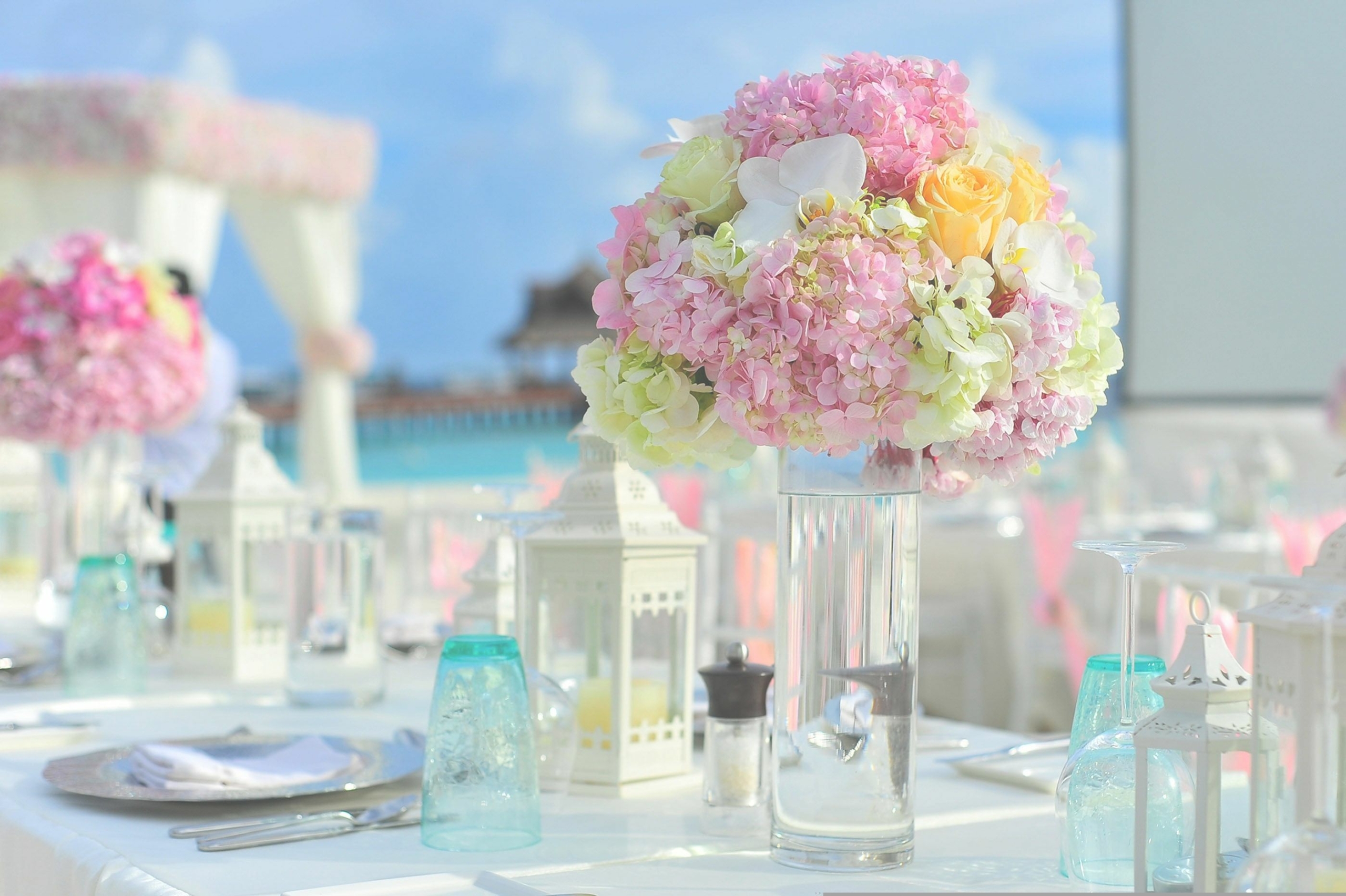 Pastel hydrangea bouquet (pink, cream, yellow) in a tall glass vase on a white-draped wedding table centerpiece