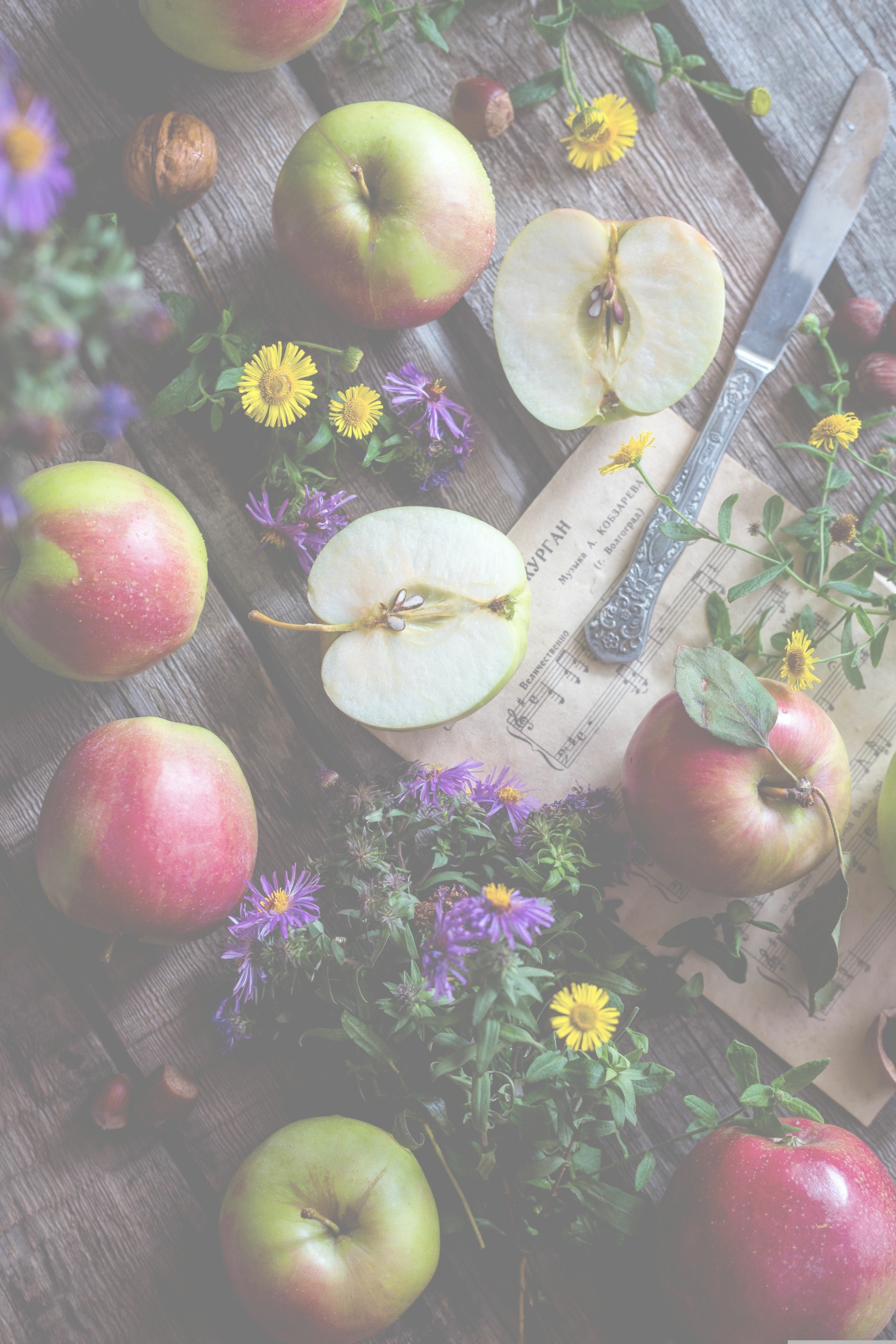 Rustic wooden table with whole and halved apples, a knife, and bright yellow and purple flowers scattered around. Tucked among them is an old newspaper.